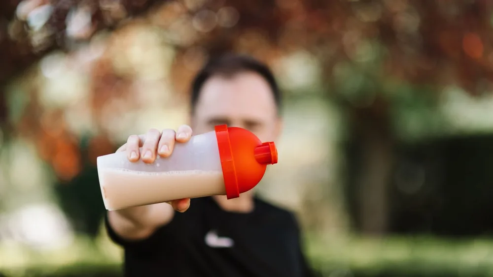 Focus on a man holding a plastic protein shake tumbler outdoors with a blurred background.
