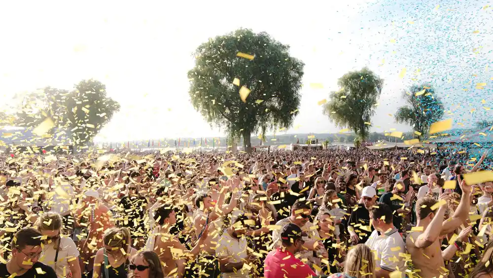 A lively crowd enjoying an outdoor music festival with confetti under a sunny sky.