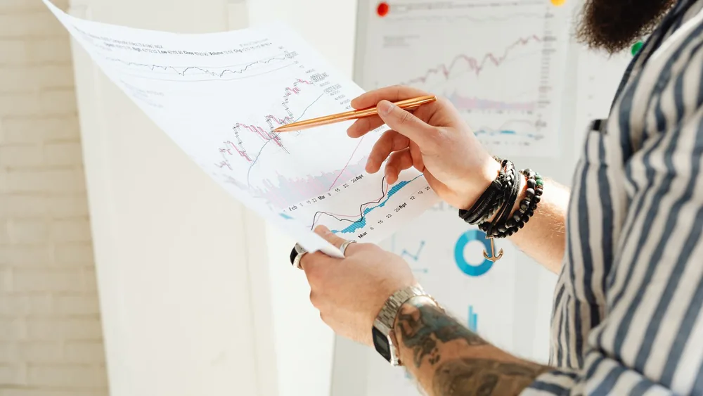 A man analyzing stock market charts with a pen, holding a paper report indoors.