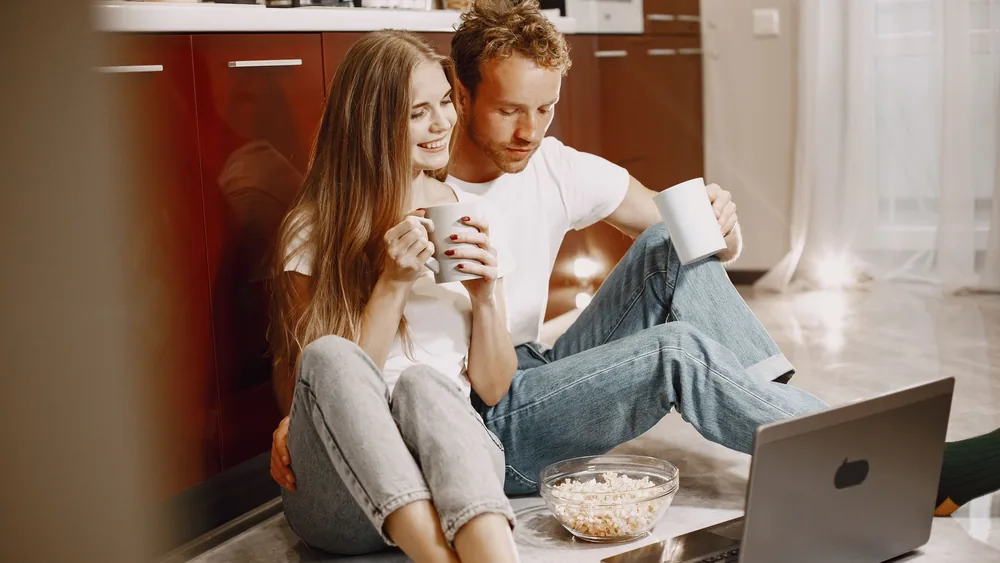 Young couple enjoying a cozy moment with popcorn and coffee by the computer at home.