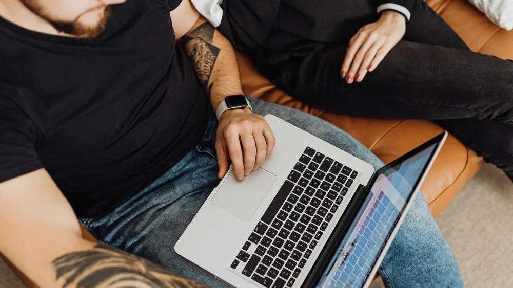 A man with tattoos browsing a laptop while sitting comfortably on a sofa indoors.