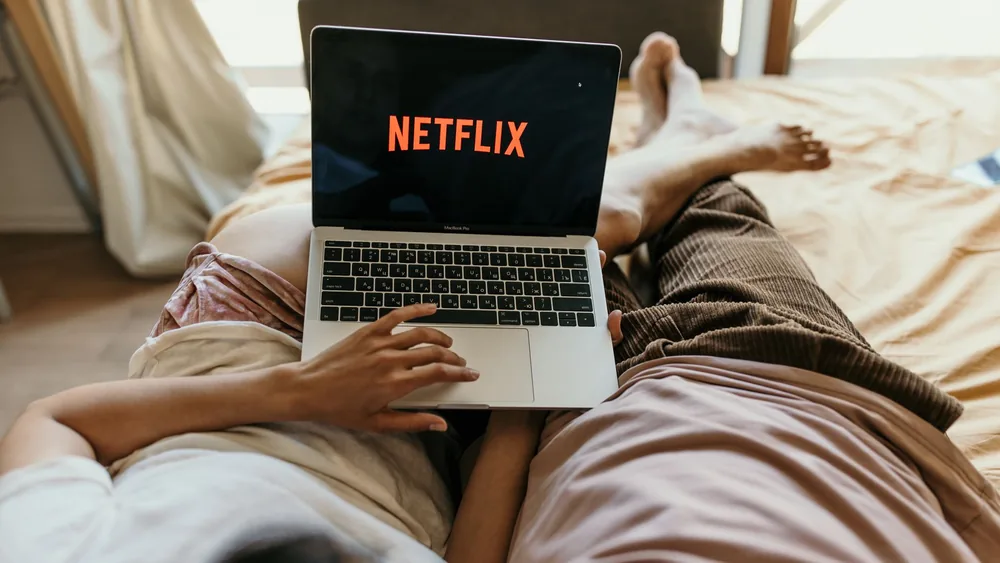 A couple lying comfortably in bed watching Netflix on a laptop, enjoying leisure time together indoors.