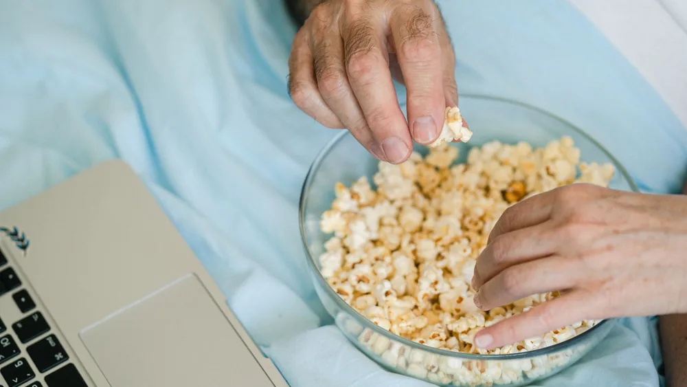 Hands reach for popcorn in a glass bowl next to a laptop, set for a cozy movie night at home.