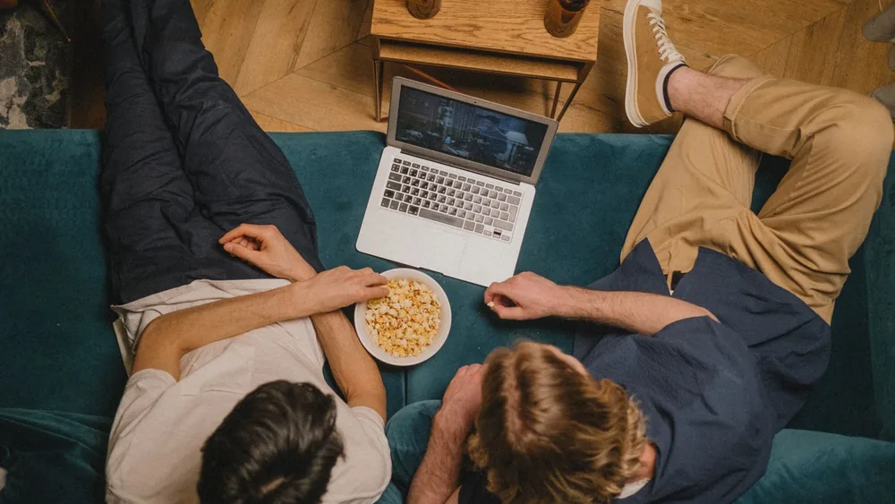 Two friends enjoying a movie on a laptop with popcorn from a cozy couch perspective.