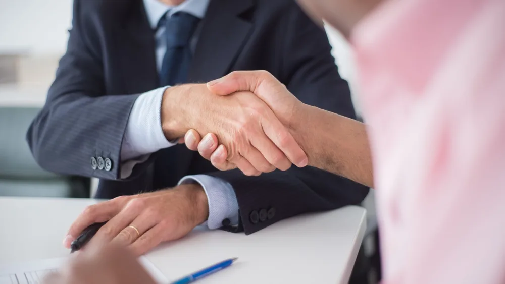 Two businessmen shaking hands across table, symbolizing agreement and partnership in an office environment.