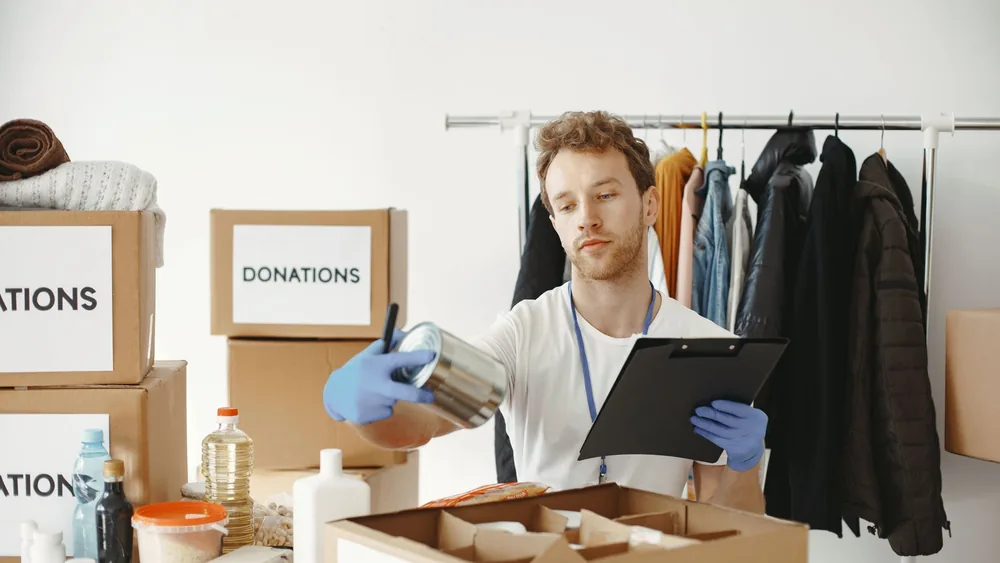 A volunteer organizes food and clothing donations in a community center, maintaining records on a clipboard.