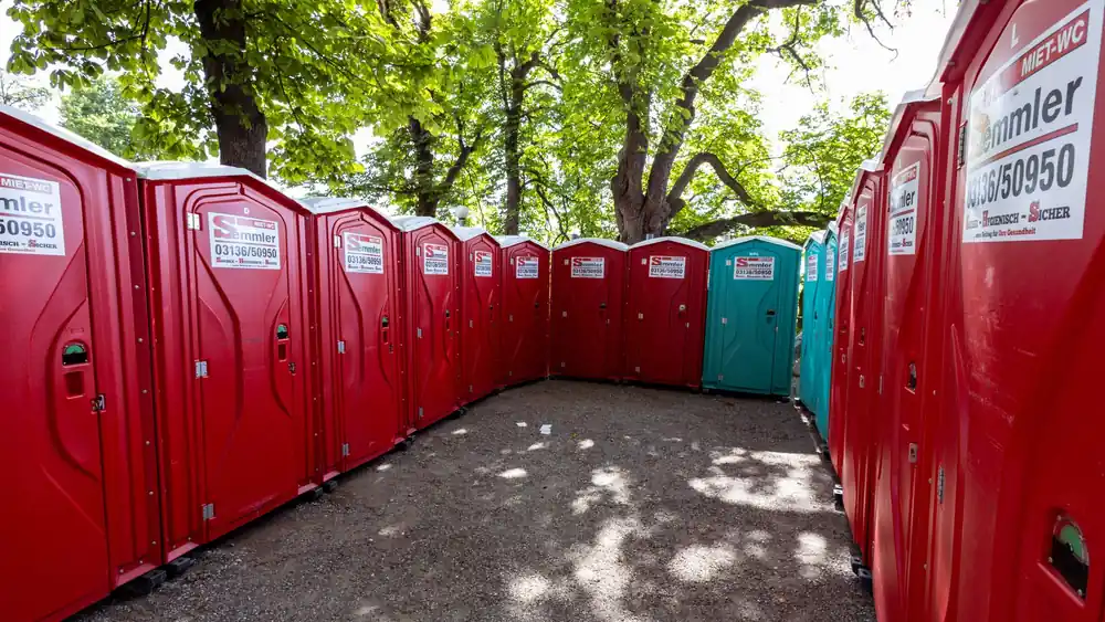 A line of red portable toilets under green trees in a sunny outdoor setting.