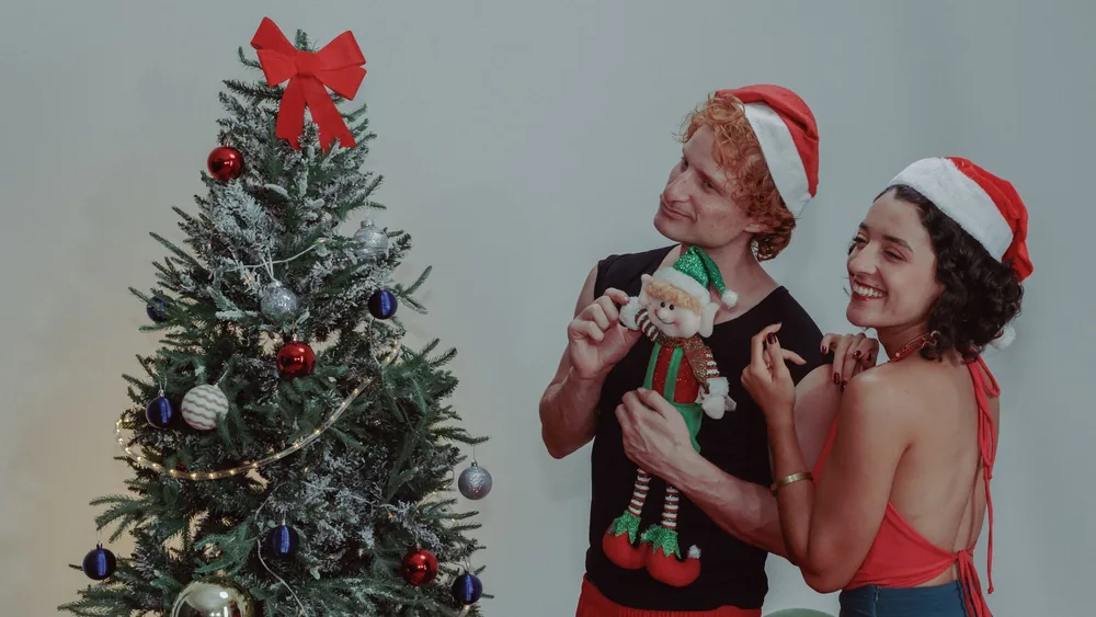 A happy couple in Santa hats decorating a Christmas tree in festive attire indoors.
