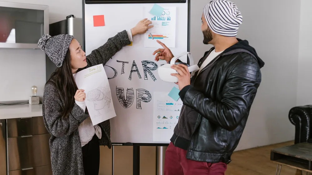 Two young adults presenting startup business ideas on a flipchart indoors.