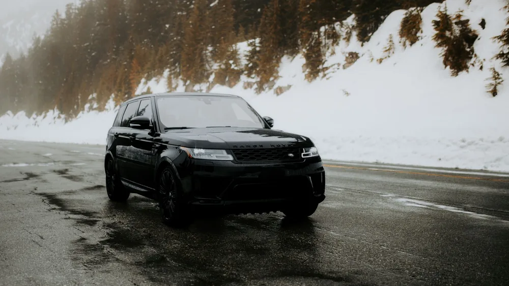 A sleek black SUV navigates a wet, snow-covered mountain road surrounded by evergreen trees.