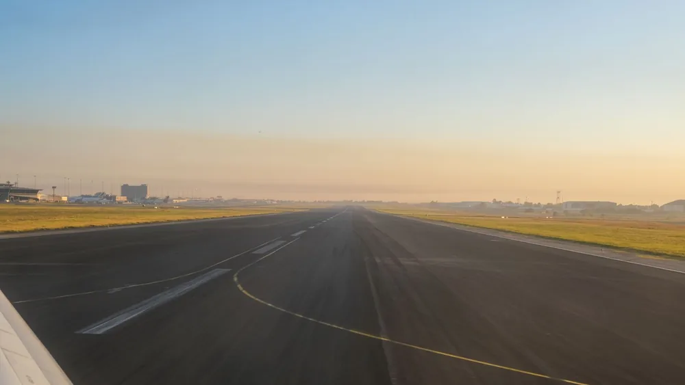 An empty airport runway at sunrise, showcasing a clear sky and distant airport buildings.