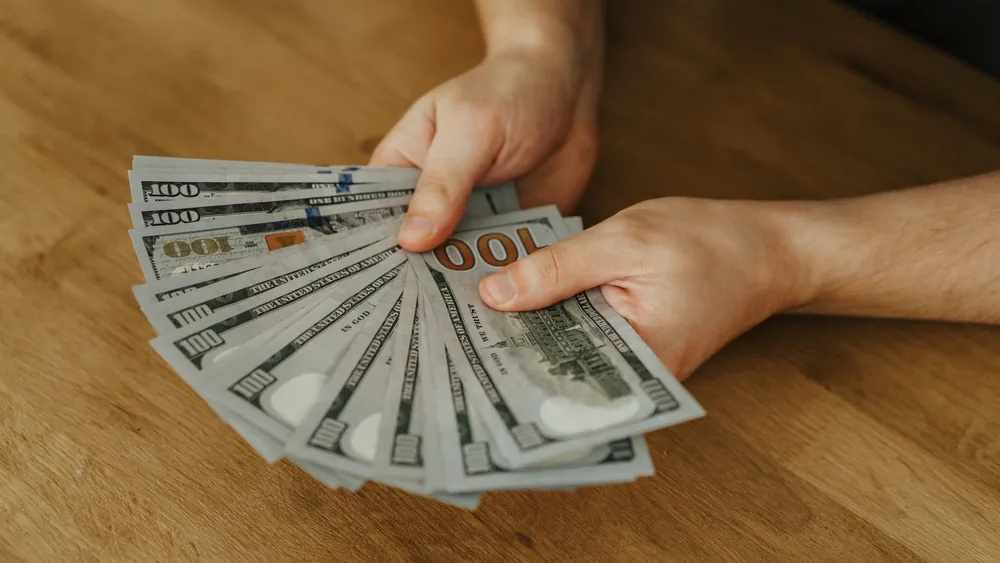 Close-up of hands holding several 100 US dollar bills on a wooden surface.