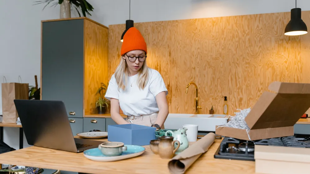Young woman packaging handmade ceramics in a modern kitchen for her online business. Warm interior setting.