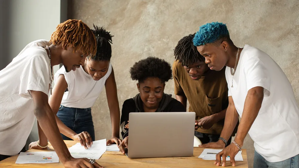 A diverse group of young adults collaborating on a laptop in an indoor workspace.