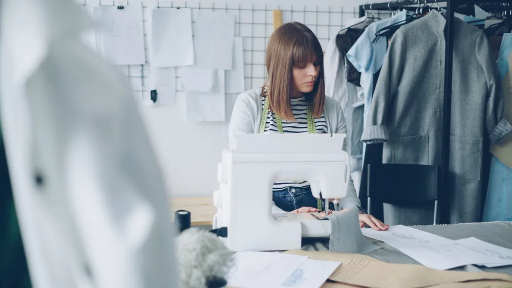 Female designer sewing in studio, surrounded by fashion sketches and fabric samples.
