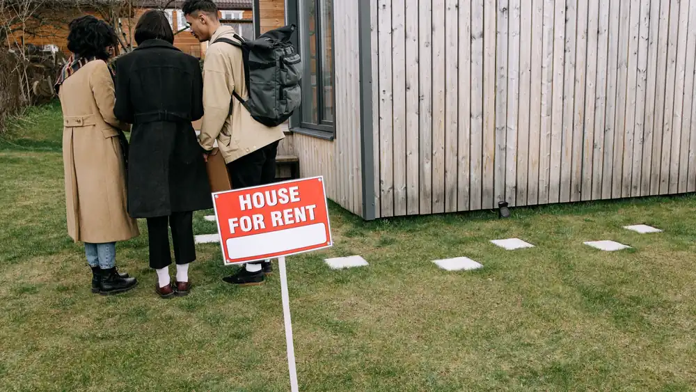 Group of adults viewing a wooden house with a 'House for Rent' sign on the lawn.