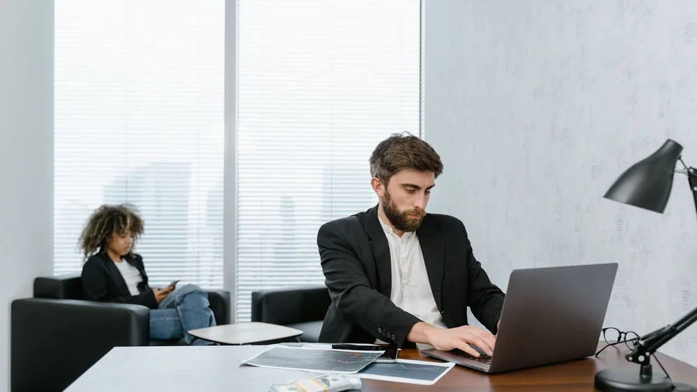 Two professionals working in an office, focused on tasks with laptops and documents.