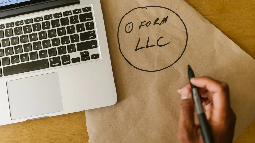 Close-up of hand writing 'Form LLC' on craft paper beside a laptop, indicating business planning.