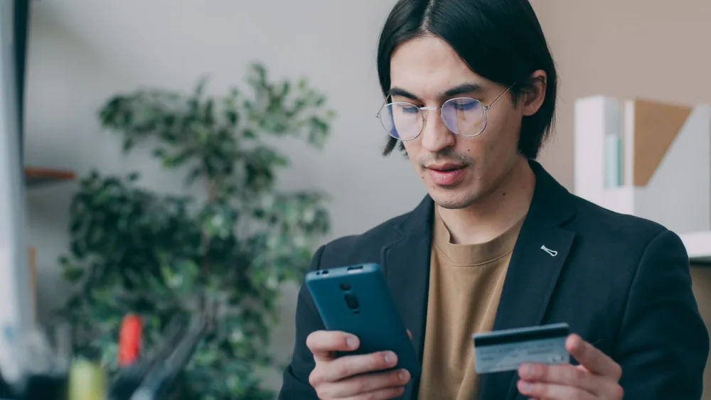 Businessman in office attire using a smartphone and holding a credit card, focusing on online payment process.