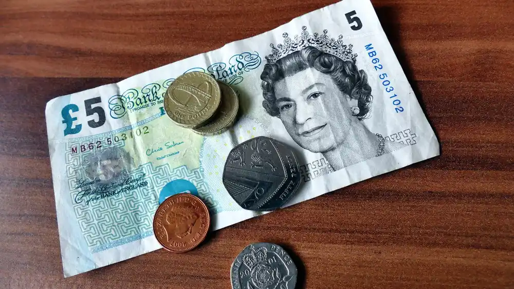 Close-up of a five-pound note and coins representing UK currency on a wooden surface.