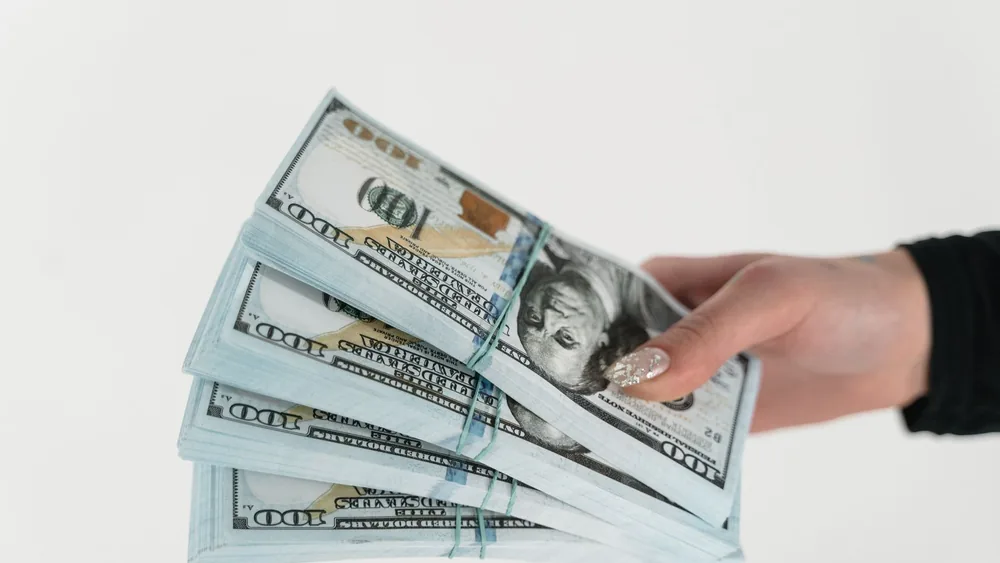 Close-up of a hand holding several stacks of US hundred dollar bills on a white background.