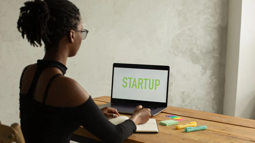 Back view of anonymous young African American businesswoman with bare shoulder and eyeglasses sitting at table with netbook with startup on screen and writing notes in copybook