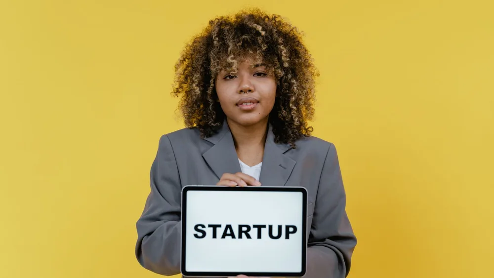 Portrait of a woman holding a tablet showing the word 'STARTUP' against a yellow background.