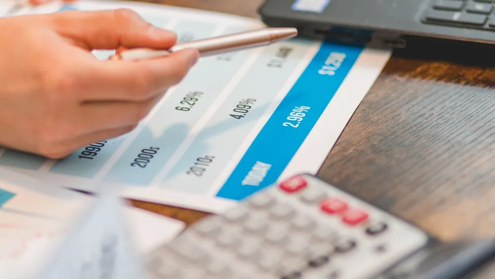 Close-up of a hand using a ballpen and calculator to analyze interest rates on a chart.