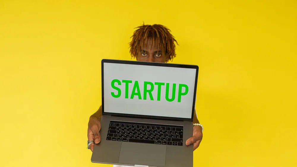 A young black man holds a laptop displaying 'Startup' against a vibrant yellow background.