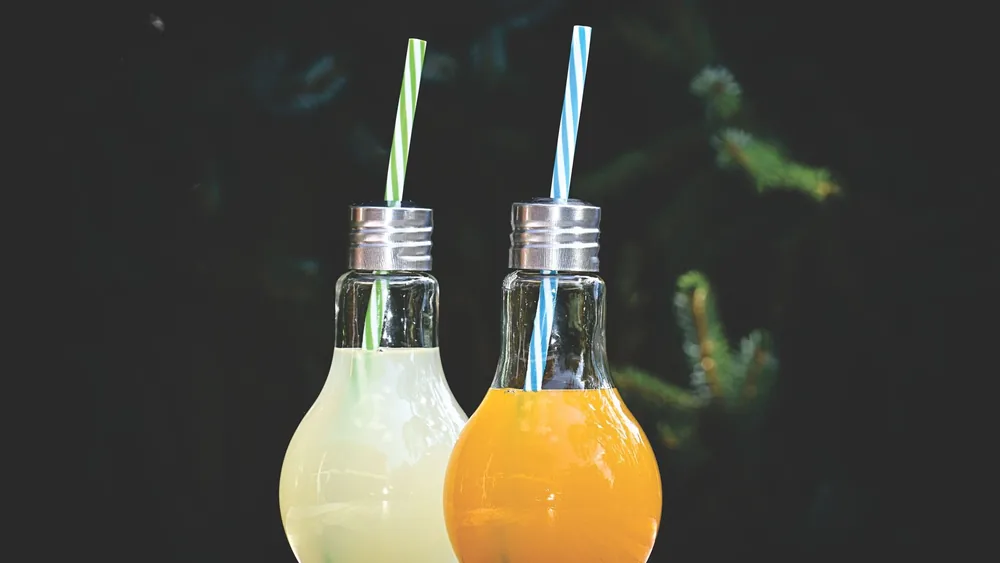 Two vibrant fruit juices in lightbulb-shaped glasses with straws on a colorful table.