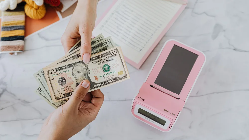 Close-up of hands exchanging US dollars with a pink calculator on a marble surface.