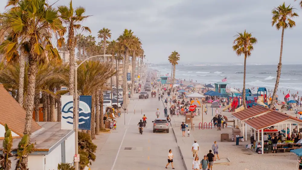 Bustling beach and boardwalk with palm trees, people, and ocean views during sunset.