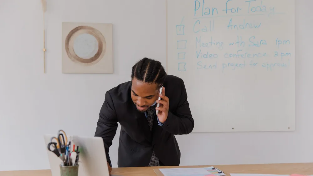 Businessman in an office making a call while working on a laptop with office supplies.