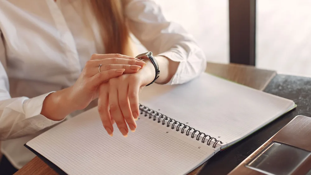 Unrecognizable crop female entrepreneur in formal clothes sitting at wooden table near laptop with copybook and checking time on wristwatch