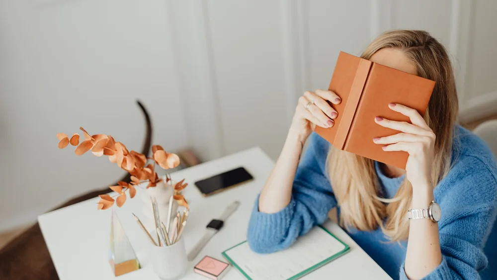 A woman in a blue sweater reading a book at a modern workspace indoors.