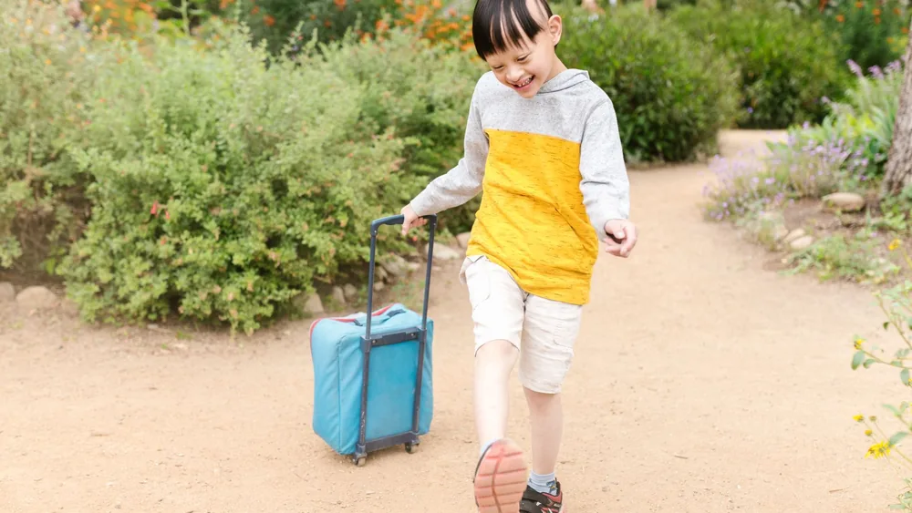 Happy child pulling a blue suitcase in a garden, enjoying a sunny day outdoors.