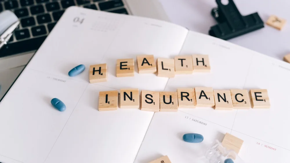 Scrabble tiles spelling 'Health Insurance' on planner with pills and laptop, symbolizing healthcare planning.