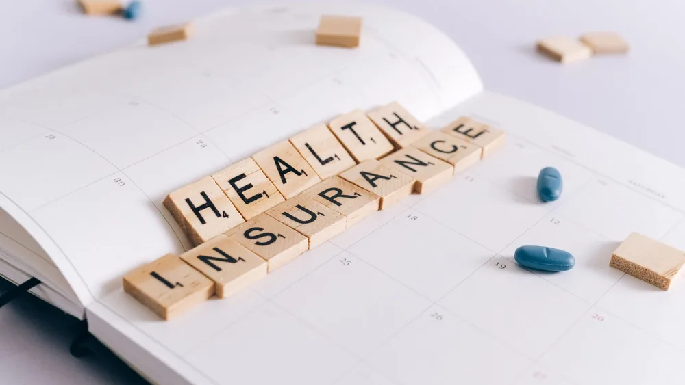 Scrabble tiles spelling 'Health Insurance' on a calendar with pills.