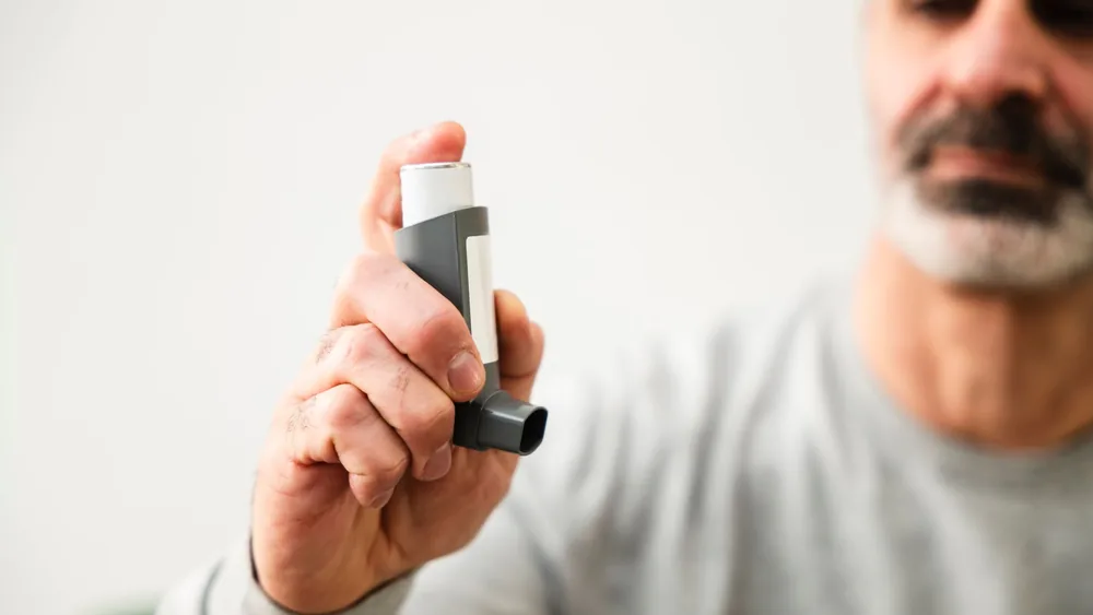 Middle-aged man using an inhaler to manage respiratory issues indoors.