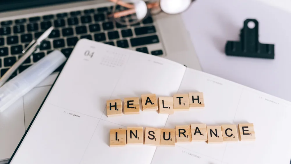 Close-up of Scrabble tiles spelling 'Health Insurance' on a planner with a laptop.