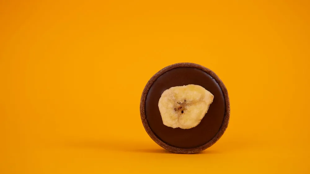 A minimalistic shot of a chocolate tart topped with banana slice on a bold yellow background.