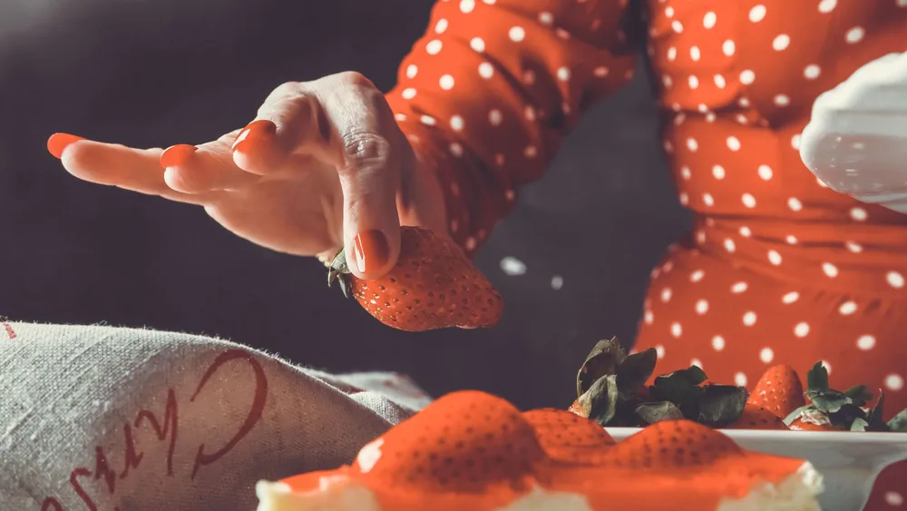 Woman in red polka dot dress placing fresh strawberry on cheesecake slice.