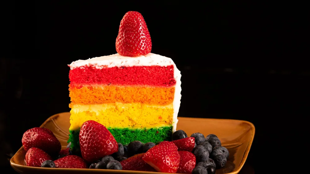 Vibrant rainbow cake slice topped with strawberries and blueberries on a plate.