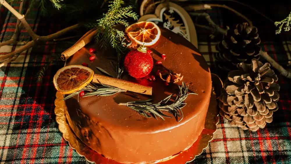 Top-down view of a chocolate cake decorated with cinnamon, orange slices, pine cones, and festive ornaments.