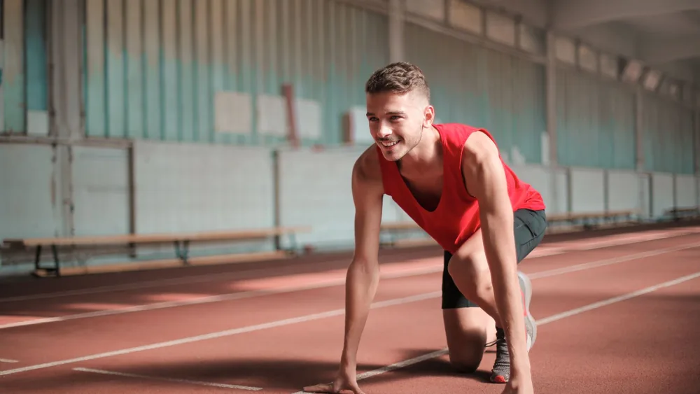 A young male athlete preparing to start running on an indoor track with a smile.