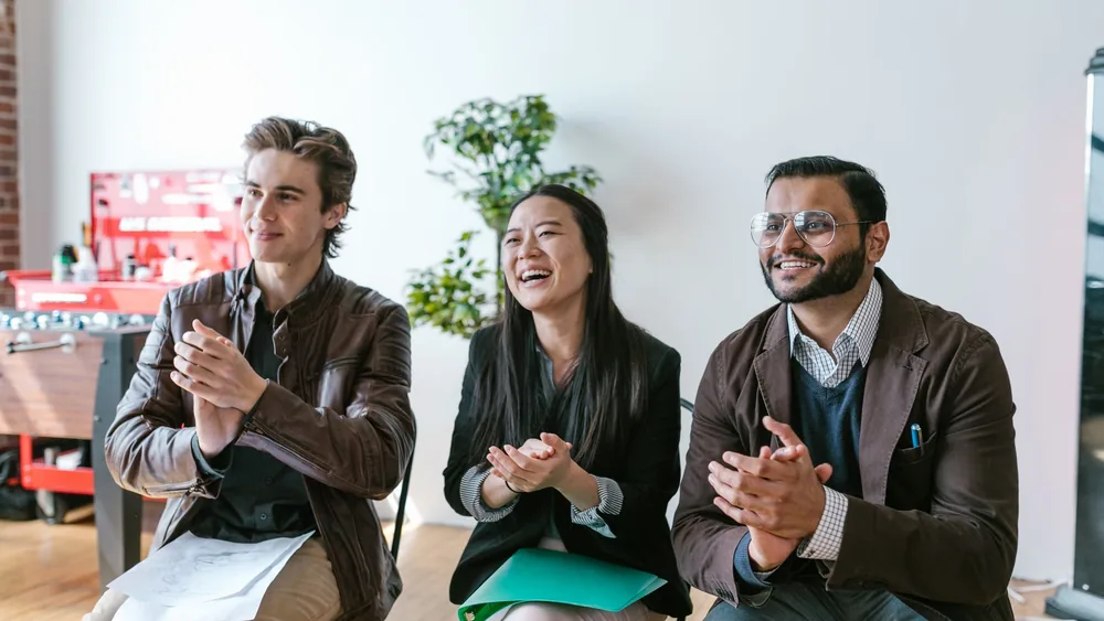 Three diverse team members clapping and smiling during an indoor meeting, showing positivity and teamwork.