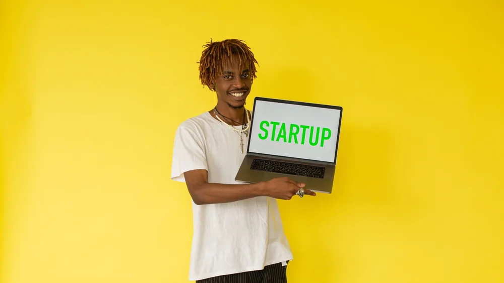 Smiling man holding a laptop with 'STARTUP' on screen against vibrant yellow background.
