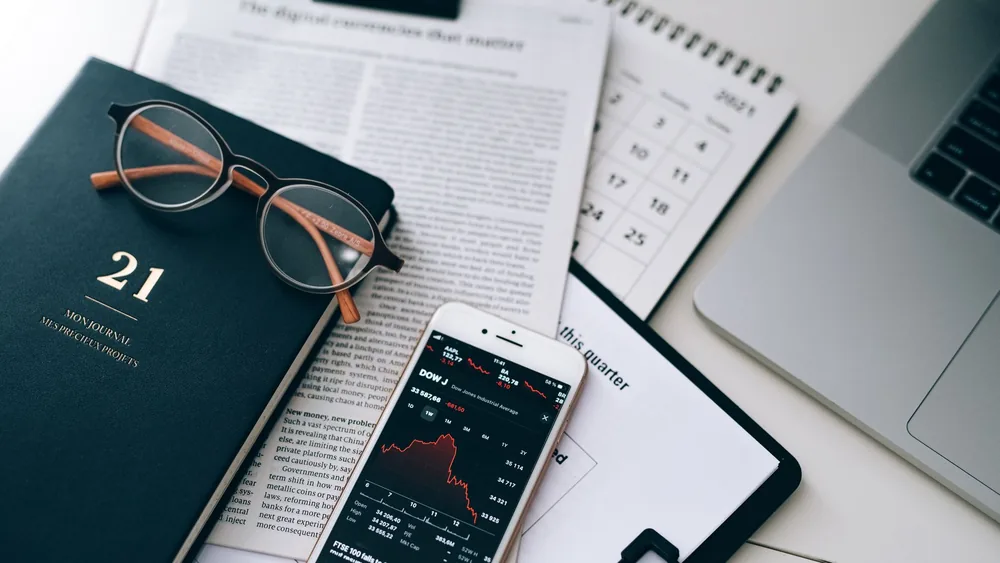 A stylish workspace featuring financial documents, eyeglasses, an iPhone displaying stock data, and a laptop.