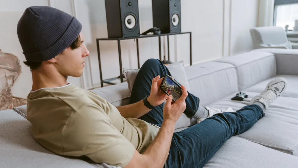 Casual young man playing mobile games on sofa with speakers in the background.