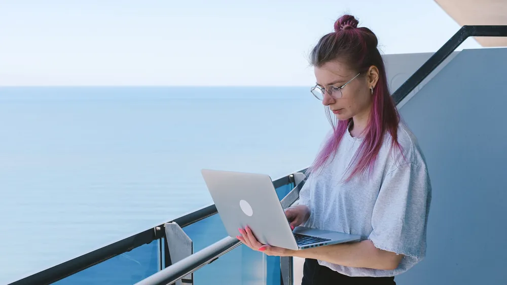 A young woman works remotely on her laptop by the sea on a sunny day, embodying the digital nomad lifestyle.
